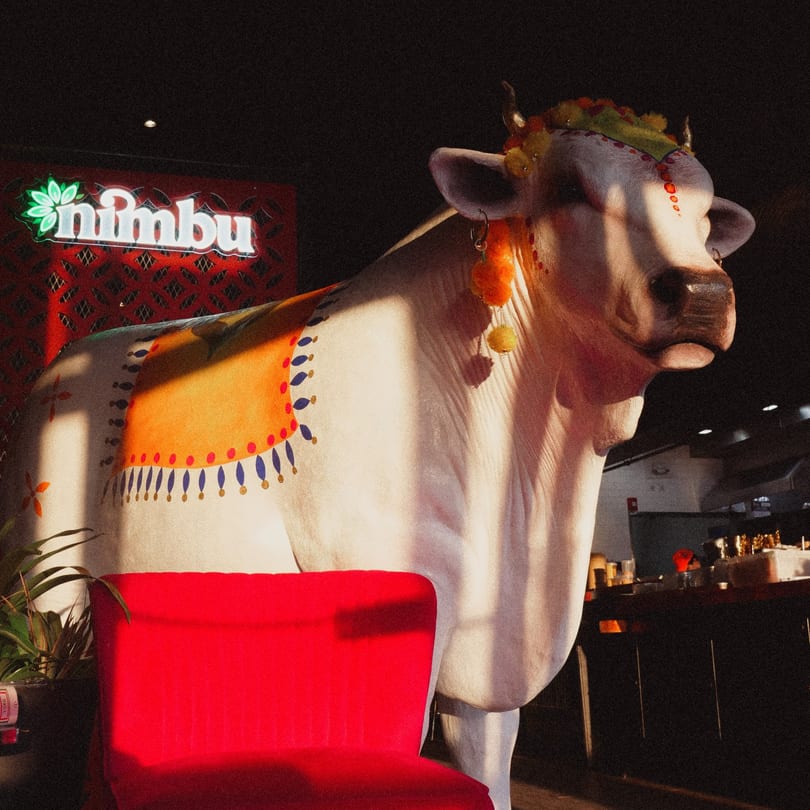 A large, decorated white cow statue stands next to a red chair, with the Nimbu logo in the background.